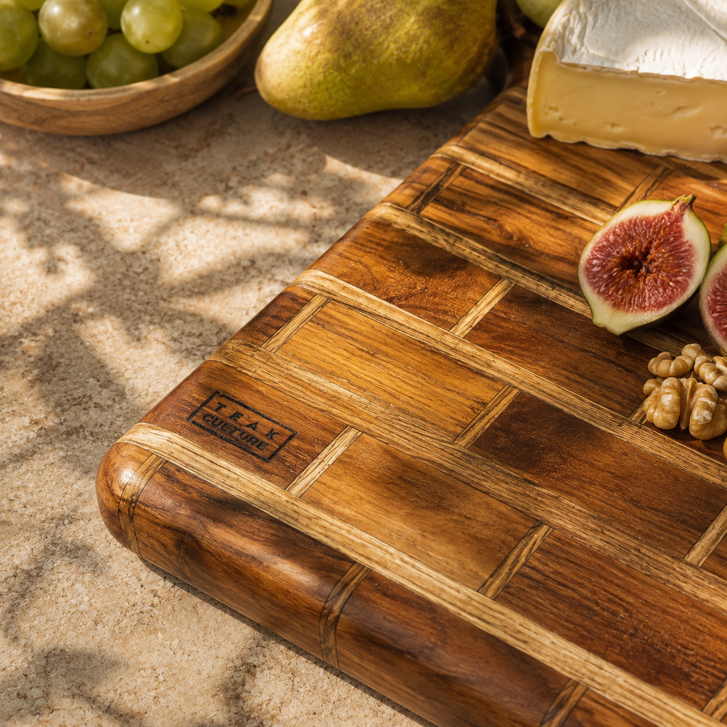 A rectangular wooden cutting board with a basketweave pattern, shown on a stone surface with food props including grapes, a pear, figs, walnuts, and a wedge of cheese. The board has a visible 'TRAK TABLES CO.