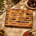 A rectangular wooden cutting board with a brick-like pattern of warm and dark wood tones, styled on a stone surface with food props including grapes, figs, olives, cheese, and sliced salami.