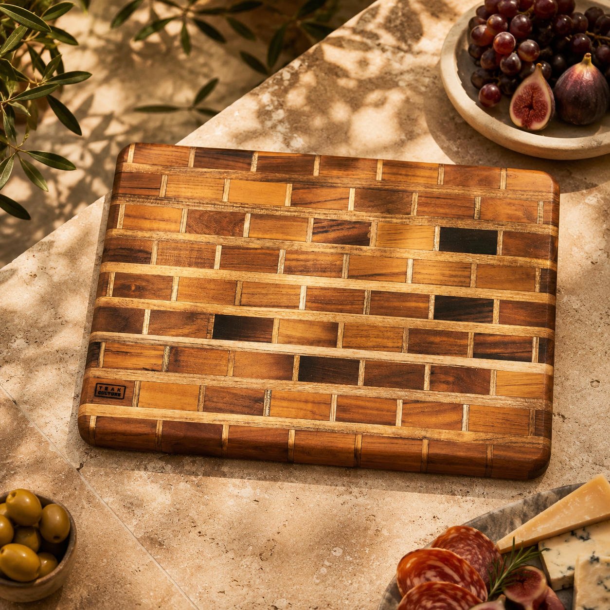 A rectangular wooden cutting board with a brick-like pattern of warm and dark wood tones, styled on a stone surface with food props including grapes, figs, olives, cheese, and sliced salami.