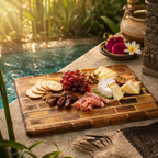 A rectangular wooden charcuterie board with a woven basket-style border, styled with crackers, red grapes, dates, rolled salami, cheese wedges, and nuts, set on a stone patio beside a pool with warm sunlight and tropical greenery in the background.