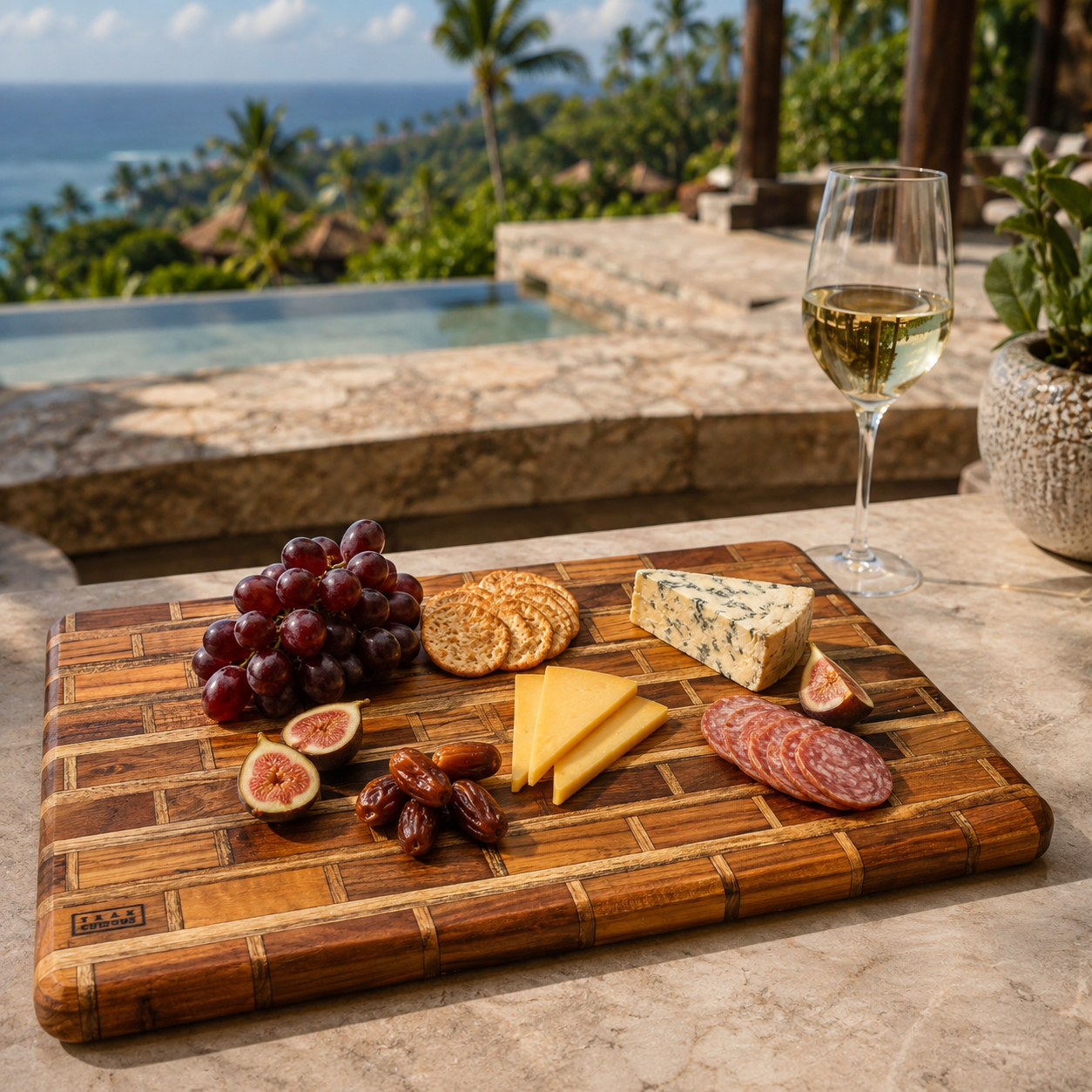 A rectangular bamboo charcuterie board with a basket-weave edge design, styled with cheese, crackers, grapes, figs, dates, and salami on a stone patio next to a glass of white wine, with a tropical ocean and pool in the background.